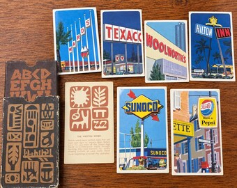 May include: A collection of vintage playing cards featuring illustrations of Wards, Texaco, Woolworths, Hilton Inn, Sunoco, and Pepsi signage, alongside a brown card box with Egyptian-style hieroglyphic patterns and alphabet letters.