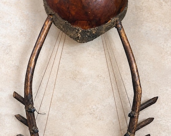 May include: A stringed musical instrument featuring a large, round, brown gourd resonator attached to two curved wooden arms. Each arm has three wooden pegs with thin strings stretched across the frame. A sticker reads HAND CARVED MADE IN KENYA.