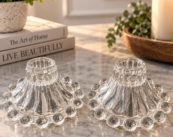 May include: A pair of clear glass candle holders featuring a wide, fluted base adorned with a decorative border of small, spherical glass beads around the bottom edge. Each holder has a central opening designed to support a single taper candle.