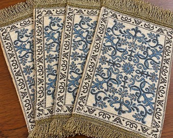 May include: A set of four rectangular fabric placemats featuring a cream-coloured base with an intricate blue floral and scrollwork pattern. Each placemat is bordered by a decorative frame and finished with metallic gold fringe on the top and bottom edges.