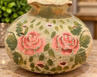 May include: A round, weathered ceramic vase with a scalloped rim, featuring a pale green base colour decorated with hand-painted pink roses and dark green leaves. The surface has a textured, aged appearance with visible signs of wear.