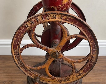 May include: A vintage cast iron hand-cranked grain mill with a distressed red and yellow paint finish. The circular wheel features the embossed text NATIONAL and includes a wooden handle. A large hopper sits at the top of the metal frame.