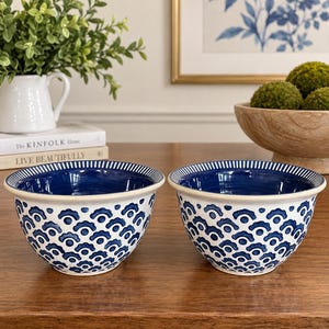 May include: Two ceramic bowls featuring a white exterior with a repeating blue scalloped pattern and a solid deep blue interior. The rims are decorated with a thin blue and white striped design. The bowls have a smooth, rounded shape and a glossy finish.