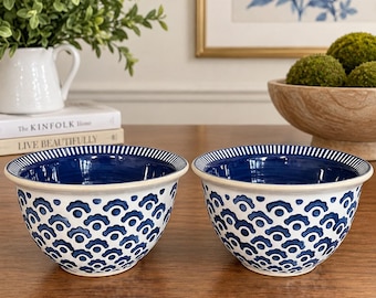 May include: Two ceramic bowls featuring a white exterior with a repeating blue scalloped pattern and a solid deep blue interior. The rims are decorated with a thin blue and white striped design. The bowls have a smooth, rounded shape and a glossy finish.
