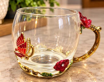 May include: A clear glass mug featuring a gold-toned metal base and handle decorated with red enamel flowers and a red butterfly with small clear crystals. The butterfly is attached to the side of the glass, while the handle is shaped like a stem.