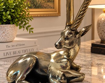 May include: A polished brass sculpture of a gazelle in a resting position. The animal is depicted with its legs tucked underneath its body and features two long, curved, spiralled horns extending upwards from the top of its head.