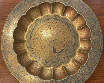 May include: A round brass decorative plate featuring an etched peacock design in the centre. The plate has a scalloped inner rim with alternating gold and red floral patterns, surrounded by a wide outer border with intricate blue floral engravings.