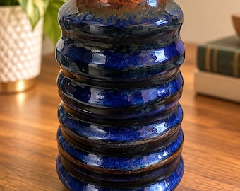 May include: A cylindrical ceramic vase featuring a deep cobalt blue glaze with horizontal ribbed bands. The rim and upper section transition into a warm, metallic copper and bronze tone, creating a contrast against the glossy blue body.
