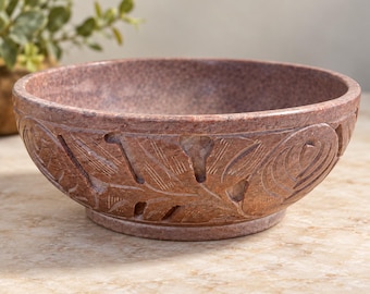 May include: A round, reddish-brown stone bowl featuring a carved exterior pattern of stylised leaves and floral shapes. The bowl has a smooth, polished interior surface and a thick, sturdy base, showcasing the natural speckled texture of the stone.