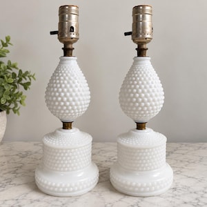 May include: A pair of white milk glass table lamps featuring a hobnail texture. Each lamp has a rounded base and a teardrop-shaped body, topped with a metallic socket. The design consists of a stacked structure with a wide bottom and a narrower top section, both covered in small, raised glass bumps.