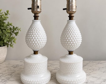 May include: A pair of white milk glass table lamps featuring a hobnail texture. Each lamp has a rounded base and a teardrop-shaped body, topped with a metallic socket. The design consists of a stacked structure with a wide bottom and a narrower top section, both covered in small, raised glass bumps.