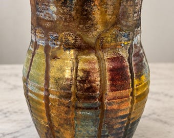 May include: A ceramic vase with a flared rim and a textured, multi-coloured glaze. The surface features vertical drip patterns in shades of metallic gold, deep brown, ochre yellow, and muted blue, creating a layered, variegated appearance.