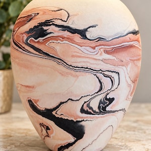 May include: A rounded vase featuring a marbled pattern with swirling colours of cream, soft peach, and deep black. The surface has a matte texture, and the vessel has a wide, curved body that tapers towards a narrow, circular opening at the top.