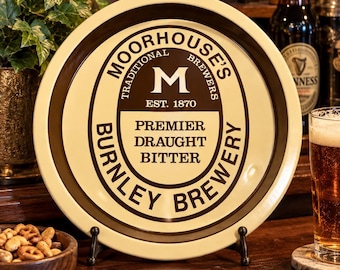 May include: A round metal serving tray with a cream-coloured background and dark brown border. The centre features the text "MOORHOUSE'S TRADITIONAL BREWERS EST. 1870 PREMIER DRAUGHT BITTER BURNLEY BREWERY" and a large letter "M" in a dark brown oval.