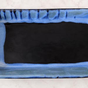 May include: A rectangular ceramic tray featuring a deep black center basin surrounded by a wide, textured border in shades of vibrant blue and dark navy. The glaze exhibits subtle variations in color intensity and vertical drip patterns.