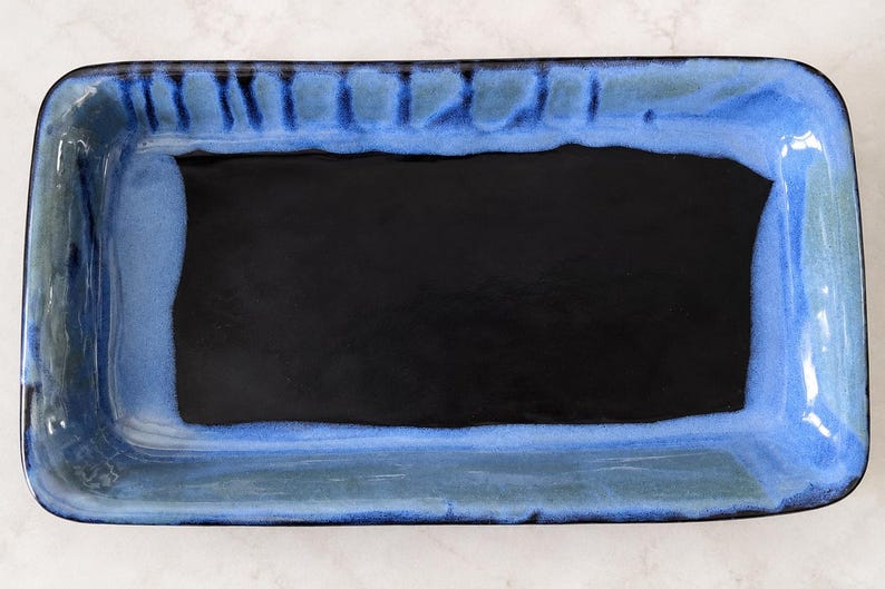 May include: A rectangular ceramic tray featuring a deep black center basin surrounded by a wide, textured border in shades of vibrant blue and dark navy. The glaze exhibits subtle variations in color intensity and vertical drip patterns.