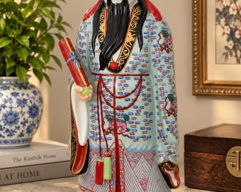 May include: A ceramic figurine of a bearded figure wearing a red hat and a light blue robe with intricate floral and geometric patterns. The figure holds a red scroll in one hand and features a red base decorated with colourful floral motifs.