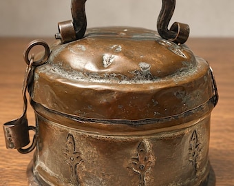 May include: A round, weathered copper container with a domed lid and a curved handle. The exterior features a hammered texture and embossed floral motifs around the base. A small metal latch mechanism is attached to the side of the vessel.