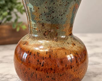May include: A ceramic vase with a bulbous base and a flared rim. The upper portion features a mottled teal and green glaze, while the lower section displays a warm, earthy brown glaze. Both sections are covered in dark brown speckles.