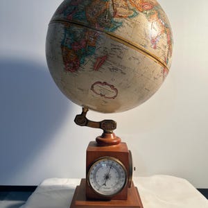 May include: A tabletop globe featuring a detailed map with muted earth tones, mounted on a wooden stand. The base includes a circular analog barometer with a white face, black markings, and a needle, integrated into the square wooden block support.
