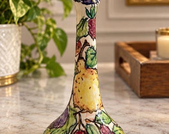 May include: A ceramic candlestick holder with a flared base and a wide top rim. The off-white surface is hand-painted with a colourful botanical pattern featuring yellow pears, clusters of purple grapes, red berries, and green leaves.