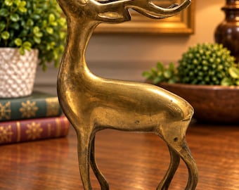 May include: A polished brass figurine of a standing deer with its head tilted upwards. The sculpture features smooth, metallic surfaces, slender legs, and curved antlers, capturing the animal in a graceful, alert pose with a warm golden tone.