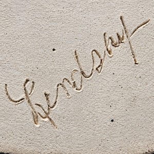 May include: A light beige, textured surface featuring the name Lindsay etched in a cursive, handwritten script. The engraving is deeply carved into the material, creating a distinct contrast between the recessed lines and the surrounding surface.