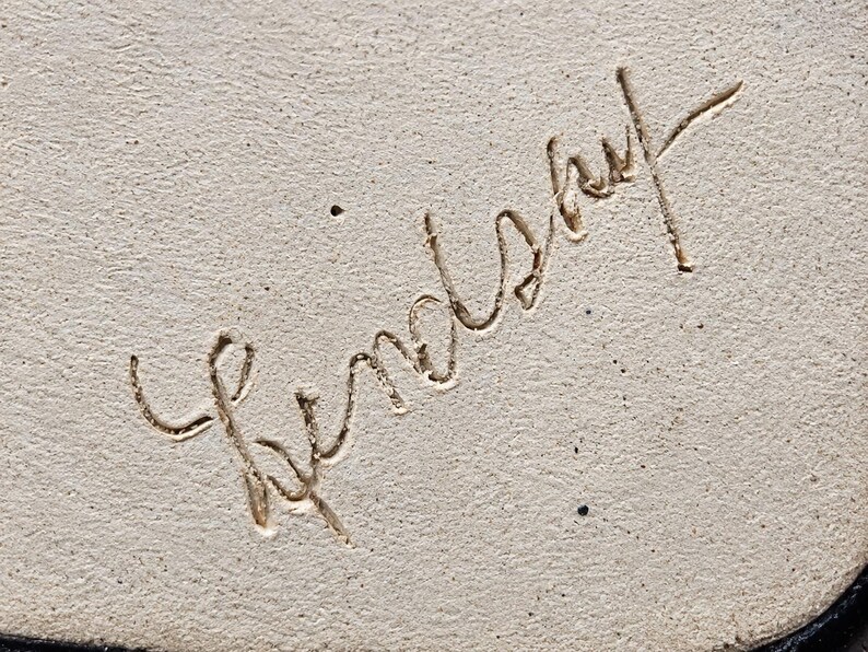 May include: A light beige, textured surface featuring the name Lindsay etched in a cursive, handwritten script. The engraving is deeply carved into the material, creating a distinct contrast between the recessed lines and the surrounding surface.
