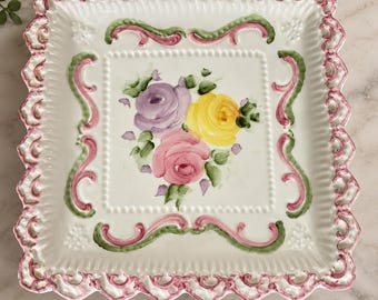May include: A square ceramic plate with a white base, featuring a central hand-painted bouquet of purple, yellow, and pink roses with green leaves. The border has a raised, scalloped edge painted in pink and green with intricate scrollwork.