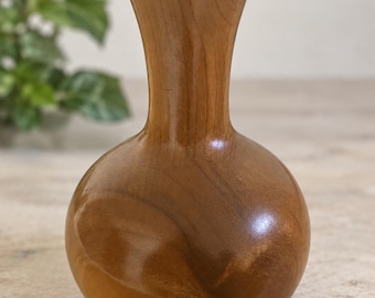 May include: A polished wooden vase with a smooth, rounded body and a slender, flared neck. The natural wood grain features warm brown tones with visible swirling patterns throughout the surface, ending in a circular opening at the top.