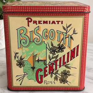 May include: A square red metal tin featuring a cream-colored label with the text Premiati Biscotti Gentilini Roma. The design includes a green illustration of a sailboat on water, dark floral branches, and a border pattern of small gold stars.