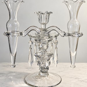 May include: A clear glass candelabra featuring a central candle holder flanked by two tall, trumpet-shaped glass vases. The structure is adorned with hanging faceted glass crystals suspended from the central section by small metal rings.