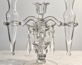 May include: A clear glass candelabra featuring a central candle holder flanked by two tall, trumpet-shaped glass vases. The structure is adorned with hanging faceted glass crystals suspended from the central section by small metal rings.