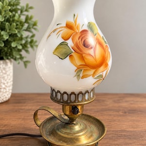 May include: A table lamp with a white milk glass shade featuring hand-painted orange and yellow roses. The base is made of aged brass with a circular tray, a small handle, and a black rotary switch. The shade has a ruffled top edge.
