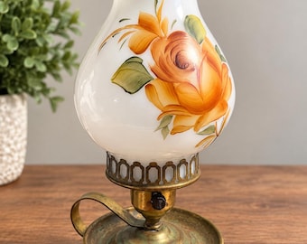 May include: A table lamp with a white milk glass shade featuring hand-painted orange and yellow roses. The base is made of aged brass with a circular tray, a small handle, and a black rotary switch. The shade has a ruffled top edge.