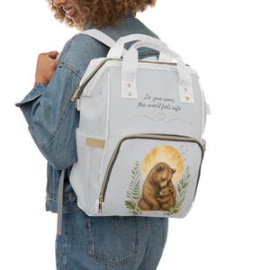 Peut inclure: Un sac à dos gris clair avec des bretelles et des bordures blanches, orné d'une illustration imprimée d'un ours brun serrant un ourson sous une lune jaune. Le texte "In your arms, the world feels safe" est imprimé en noir au-dessus.