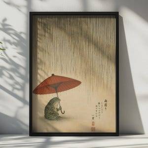以下が含まれることがあります： 垂直の雨筋の下で赤い紙の傘を差す緑色のカエルが描かれた額装アートプリント。ベージュの背景に日本語の書道と赤い印章が施されており、細い黒いフレームに収められています。