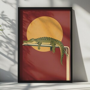 Könnte beinhalten: Ein gerahmter Wandkunstdruck, der einen grünen Alligator zeigt, der auf einem Sprungbrett vor einem tiefroten Hintergrund mit einem großen, gelben Kreis liegt, der die Sonne darstellt. Der Alligator liegt horizontal mit herabhängendem Schwanz.