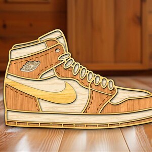 Peut inclure: Une pièce d'art mural en bois superposé en forme de basket montante. Le design présente des textures de grain de bois aux tons clairs et moyens, un swoosh latéral, un laçage frontal et un logo Air Jordan circulaire sur le panneau latéral.