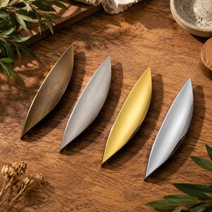 May include: Four elongated, leaf-shaped cabinet pulls arranged in a row. The pulls feature distinct finishes: a brushed antique brass, a brushed nickel, a polished gold, and a polished chrome, each with a smooth, curved, and tapered design.