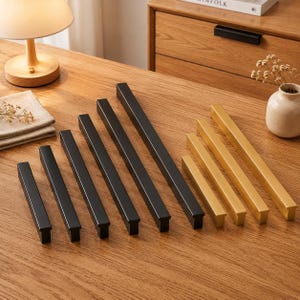 Puede incluir: Una colección de tiradores lineales para armarios dispuestos en fila, con cinco tiradores de metal negro mate de longitudes crecientes seguidos de tres tiradores de metal dorado cepillado de longitudes crecientes, todos con un perfil rectangular.