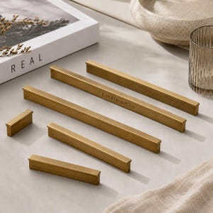May include: A collection of five brass-toned metal cabinet pulls in varying lengths, featuring a linear, rectangular profile. One of the longer pulls is engraved with the text LinkBeadCov. The hardware has a smooth, matte finish.