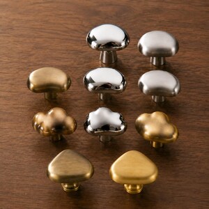 May include: A collection of ten metal cabinet knobs in various shapes and finishes. The knobs include smooth mushroom-like domes, cloud-like forms, and triangular designs in polished chrome, brushed silver, antique brass, and matte gold tones.
