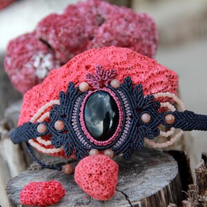 Op de afbeelding: Een zwarte macramé armband met een grote zwarte edelsteen en roze kralen. De armband ligt op een houten oppervlak met rood koraal.