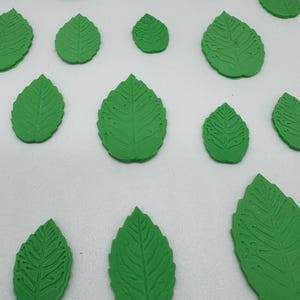 May include: A collection of small, flat, green leaf-shaped decorations arranged in a scattered pattern. Each piece features a detailed, embossed vein texture on the surface, mimicking the appearance of natural foliage in various sizes.