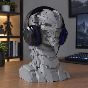 Pode incluir: Um busto de cabeça robótica cinza fosco servindo como suporte para fones de ouvido, apresentando placas mecânicas complexas e detalhes de pescoço segmentados. Fones de ouvido pretos com acentos LED circulares azuis sobre o busto.