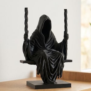 Op de afbeelding: Een matzwart beeldje van een figuur met een kap die op een schommel zit. Het figuur draagt een lang, gedrapeerd gewaad met diepe plooien en houdt twee verticale, gedraaide touwen vast die aan de zitting van de schommel zijn bevestigd. Het geheel staat op een vierkante voet.