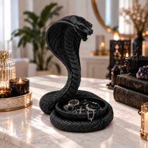 Può includere: Una scultura di cobra nero opaco con un corpo avvolto che forma una base a vassoio circolare. Il serpente è raffigurato con il cappuccio spiegato e la bocca aperta. Il vassoio contiene diversi anelli color argento, un ciondolo a croce e una luna.
