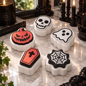 Könnte beinhalten: Ein Set aus fünf weißen Kunststoff-Drucktasten mit Halloween-Motiven: ein schwarz-weißer Totenkopf, ein weißer Geist mit schwarzen Umrissen, ein schwarzes Spinnennetz, ein orangefarbener Kürbis mit gezacktem Gesicht und ein orangefarbener Sarg.