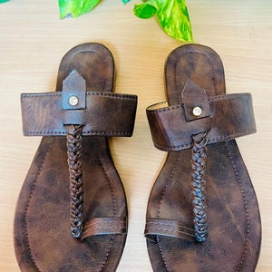 May include: A pair of dark brown flat sandals featuring a textured finish, a wide horizontal strap with a central rhinestone accent, and a braided vertical strap that connects to a toe loop. The footbed has a matching dark brown mottled pattern.